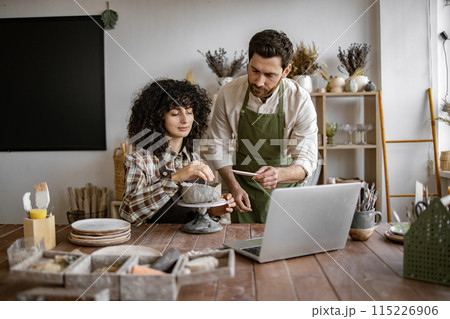 Man teaching woman pottery in studio with laptop 115226906