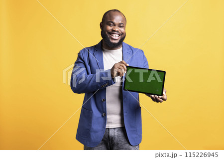 Happy african american laughing while man holding green screen tablet, doing recommendation. Overjoyed BIPOC person presenting chroma key device, isolated over studio background 115226945
