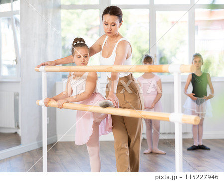Girl ballet students prepare for classes, near ballet bare stretch their leg under teacher guidance. 115227946