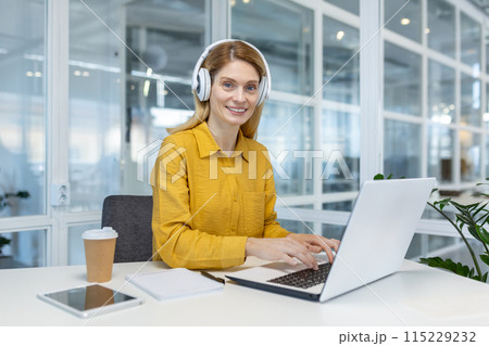 Smiling businesswoman wearing headphones working on a laptop in a modern office setting. Coffee cup and notebook are on the desk. 115229232