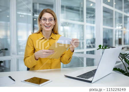 A happy woman in a yellow shirt opening an envelope at her desk in a modern office setting, expressing joy. A happy woman in a yellow shirt opening an envelope at her desk in a modern office setting, expressing joy. 115229249