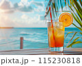 cocktail in glass with straw and ice on the table on the beach, ocean and palm leaf as background 115230818