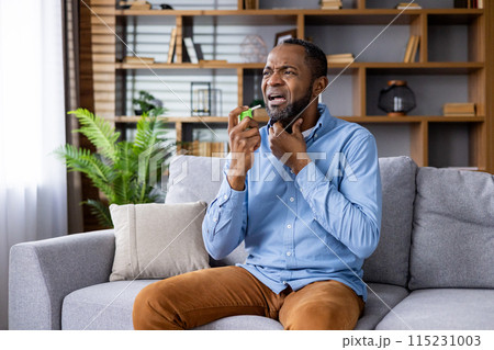 Man experiencing throat pain and using throat spray while sitting on a couch in a comfortable home setting, looking distressed 115231003