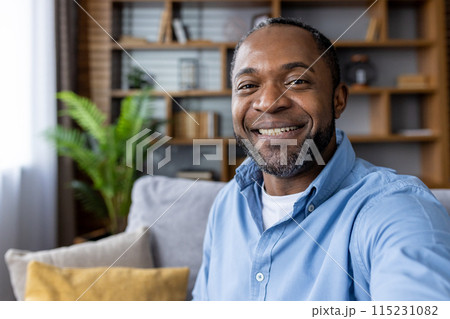 A cheerful man with a beard and blue shirt smiling warmly while sitting on a cozy couch in a modern living room 115231082