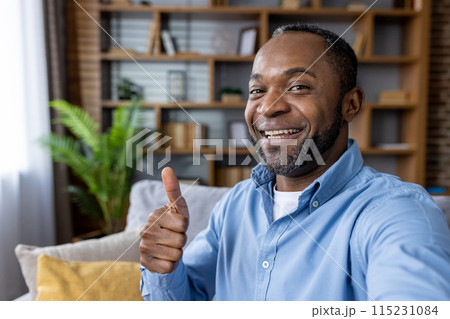 Happy man in blue shirt giving a thumbs up gesture while on a video call in a comfortable home setting. 115231084