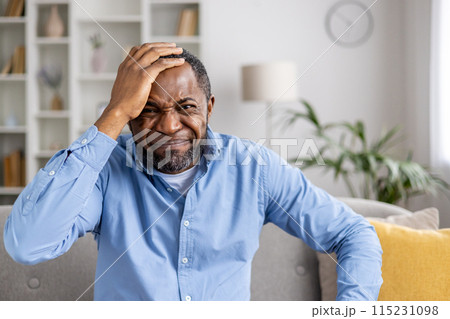 Frustrated man holding his head with a grimaced expression sitting on a couch in a living room. Emotions of confusion, stress, and frustration Frustrated man holding his head with a grimaced expression sitting on a couch in a living room. Emotions of confusion, stress, and frustration 115231098