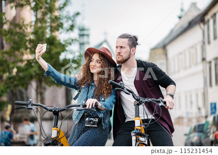 Travellers couple taking selfie, standing by electric scooters in small city, sightseeing. Young tourist using bikes during their summer trip, vacation. 115231454