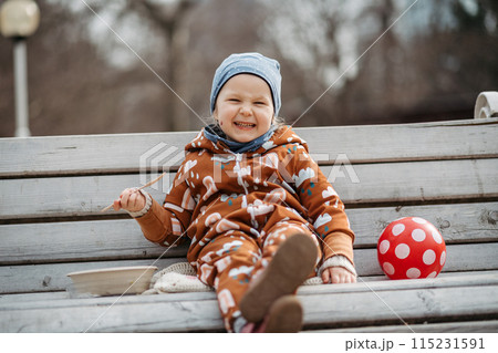 Cute toddler girl eating lunch in park, sitting on bench. Picnic in park during cold spring day. Cute toddler girl eating lunch in park, sitting on bench. Picnic in park during cold spring day. 115231591