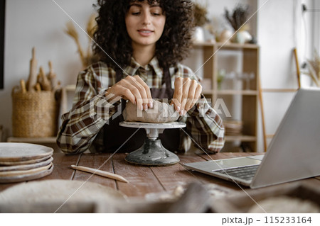 Woman creating pottery in her home studio with laptop Woman creating pottery in her home studio with laptop 115233164