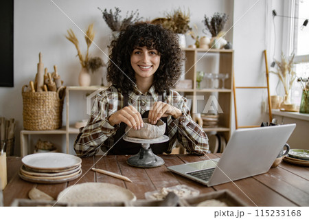 Female artist shaping pottery in studio with laptop 115233168