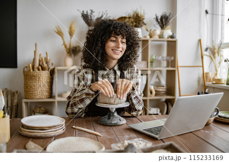 Woman shaping pottery while using laptop in studio Woman shaping pottery while using laptop in studio 115233169