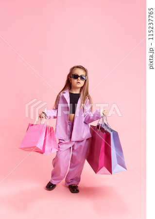 Portrait of girl dressed stylishly, holding shopping bags, dreams of future in fashion against pastel pink studio background. Portrait of girl dressed stylishly, holding shopping bags, dreams of future in fashion against pastel pink studio background. 115237765