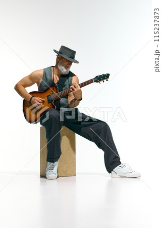 Talented man with white beard and hat captured in midst of energetic guitar performance against white studio background. Talented man with white beard and hat captured in midst of energetic guitar performance against white studio background. 115237873