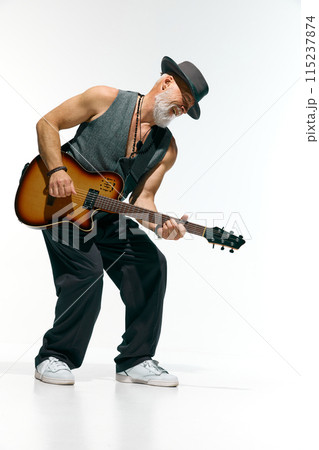 Man with beard, wearing gray sleeveless shirt and stylish hat, deeply engrossed in playing electric guitar against white studio background. Man with beard, wearing gray sleeveless shirt and stylish hat, deeply engrossed in playing electric guitar against white studio background. 115237874
