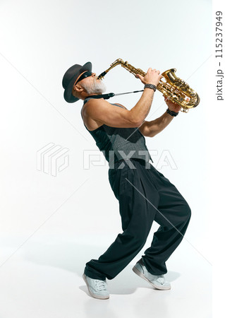 Bearded man wearing sleeveless gray vest, black pants, and stylish hat passionately plays golden saxophone against white studio background. 115237949