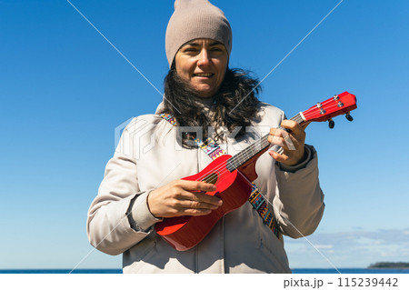 Woman playing red ukulele at the ocean edge Woman playing red ukulele at the ocean edge 115239442
