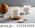 Two glasses of fresh coconut milk on wooden coasters with sliced coconut pieces and a whole coconut on a textured gray surface. 115241004