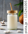 A jar of fresh coconut milk with a bamboo straw, surrounded by coconut pieces and a whole coconut on a textured surface. 115241005