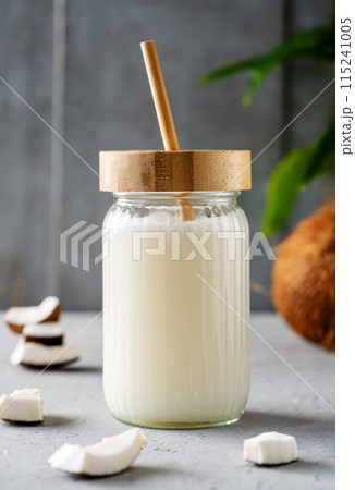 A jar of fresh coconut milk with a bamboo straw, surrounded by coconut pieces and a whole coconut on a textured surface. 115241005