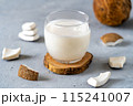A glass of fresh coconut milk placed on a wooden coaster, surrounded by coconut pieces on a textured gray surface. 115241007