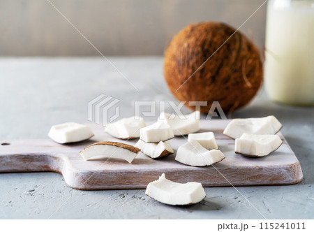 Freshly sliced coconut pieces arranged on a wooden cutting board with a whole coconut and a jar of coconut milk in the background. 115241011
