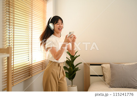 Joyful young woman holding phone listening to favorite songs and dancing in living room 115241776