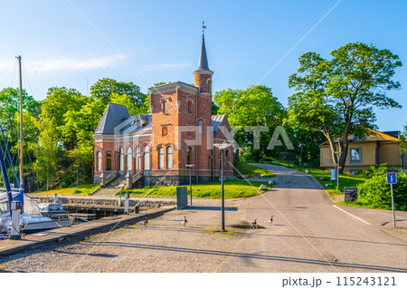A serene summer day at Skridskopaviljongen, featuring a historic building surrounded by lush greenery and a clear blue sky, with boats moored nearby and ducks crossing the path. Stockholm, Sweden 115243121
