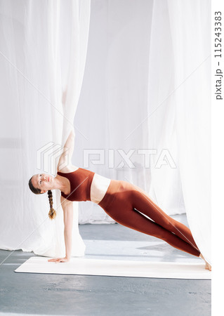 A slender woman in a brown sports uniform does vasishthasana against the background of white curtains. Yoga classes in the loft studio 115243383