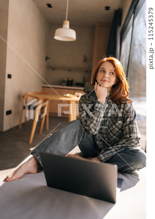 Vertical shot of positive charming young redhead woman dressed in casual clothes relaxing on wide windowsill with portable computer, shopping online, looking away with cute cheerful smile 115245379