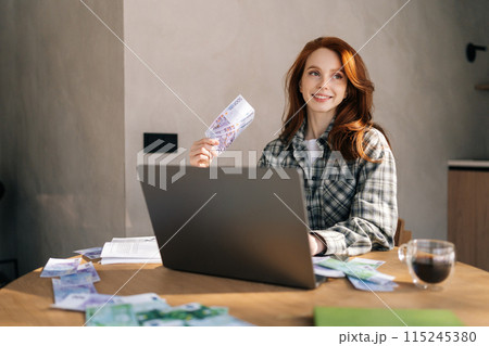 Medium shot of lucky young businesswoman holding fan of cash money euro banknotes celebrate, dance, success career, lottery jackpot game winner, big income, wealth at home office workplace desk. 115245380