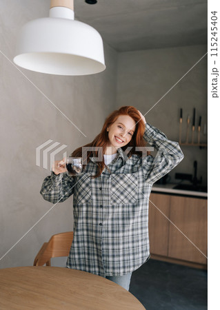 Vertical portrait of cheerful young woman enjoying holding cup drinking coffee standing in kitchen on sunny morning, smiling looking at camera. Happy female relaxing on caffeine break alone. 115245404