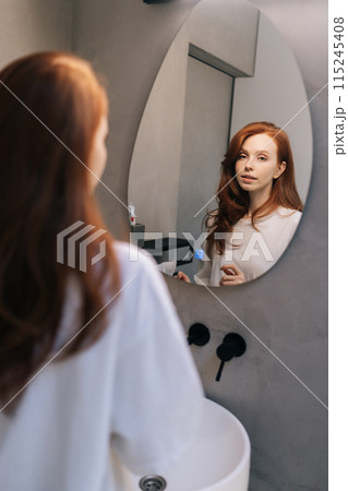 Vertical portrait of charming woman in casual clothes, preening, do morning routine, touch her hair standing in front of mirror, smiling admire herself feels satisfied with appearance and complexion. 115245408