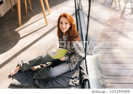 Top view of cheerful young woman holding reading book sitting by window on sunny day, smiling looking at camera, enjoying leisure activity, carefree weekend with favorite literature and home pastime. Top view of cheerful young woman holding reading book sitting by window on sunny day, smiling looking at camera, enjoying leisure activity, carefree weekend with favorite literature and home pastime. 115245409