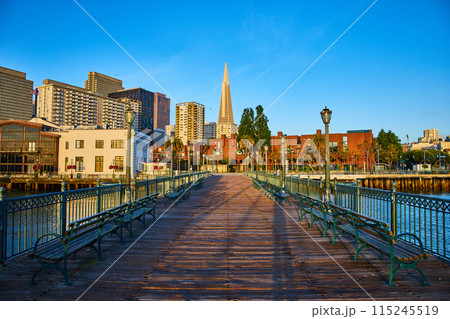 Dark wood pier at sunrise with light leading up to golden skyscrapers and blue sky in San Francisco 115245519