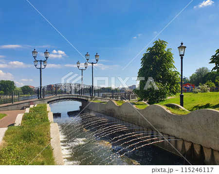 Empty small pedestrian stone bridge over water canal with fountain streams on embankment of Upper Lake - artificial city pond in Kaliningrad, Russia at sunny summer day. With no people cityscape. Empty small pedestrian stone bridge over water canal with fountain streams on embankment of Upper Lake - artificial city pond in Kaliningrad, Russia at sunny summer day. With no people cityscape. 115246117