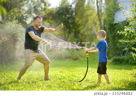 Funny little boy with his father playing with garden hose in sunny backyard. Preschooler child having fun with spray of water. Summer outdoors activity for kids. 115246882
