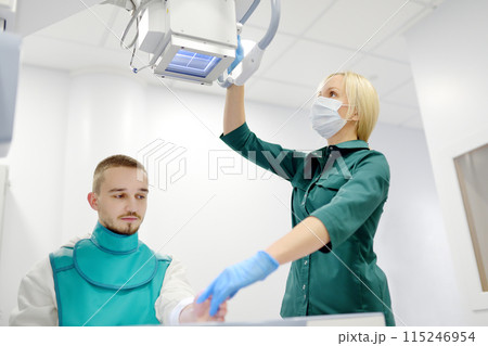Female radiologist going to made x-ray shot of young man hand in x-ray room in modern clinic. Patient wearing in protect lead apron Female radiologist going to made x-ray shot of young man hand in x-ray room in modern clinic. Patient wearing in protect lead apron 115246954