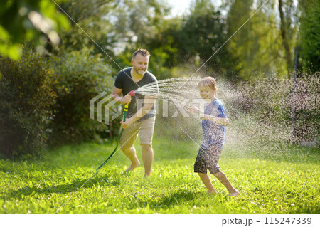Funny little boy with his father playing with garden hose in sunny backyard. Preschooler child having fun with spray of water. Summer outdoors activity for kids. Funny little boy with his father playing with garden hose in sunny backyard. Preschooler child having fun with spray of water. Summer outdoors activity for kids. 115247339