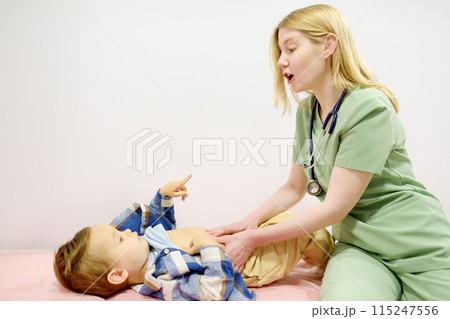 Cute toddler boy is at an appointment with pediatrician. Doctor gastroenterologist palpating belly of little child patient lying on couch in medical office. Gastroenterological diseases of kids 115247556