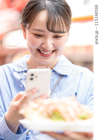 明るい屋外でたこ焼きをスマホで撮影する若くて可愛いSNS好きな女性｜大阪府大阪市道頓堀商店街 115248588