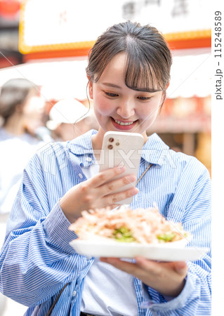 明るい屋外でたこ焼きをスマホで撮影する若くて可愛いSNS好きな女性|大阪府大阪市道頓堀商店街 明るい屋外でたこ焼きをスマホで撮影する若くて可愛いSNS好きな女性|大阪府大阪市道頓堀商店街 115248589