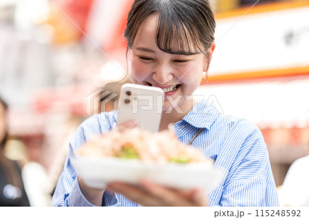 明るい屋外でたこ焼きをスマホで撮影する若くて可愛いSNS好きな女性|大阪府大阪市道頓堀商店街 明るい屋外でたこ焼きをスマホで撮影する若くて可愛いSNS好きな女性|大阪府大阪市道頓堀商店街 115248592