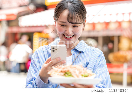 明るい屋外でたこ焼きをスマホで撮影する若くて可愛いSNS好きな女性｜大阪府大阪市道頓堀商店街 115248594