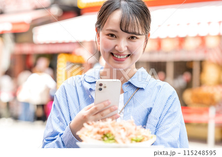 明るい屋外でたこ焼きをスマホで撮影する若くて可愛いSNS好きな女性｜大阪府大阪市道頓堀商店街 115248595