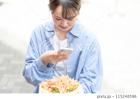 明るい屋外でたこ焼きをスマホで撮影する若くて可愛いSNS好きな女性｜大阪府大阪市道頓堀商店街 115248596