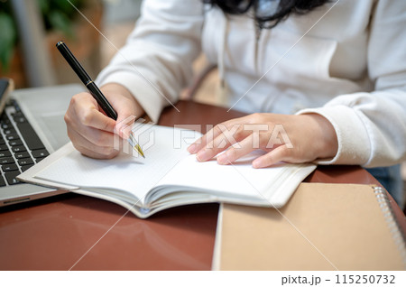 A woman is writing something in her notebook while watching a video on her laptop. 115250732