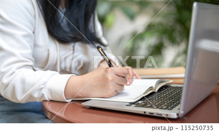 A woman is writing something in her notebook while watching a video on her laptop. 115250735