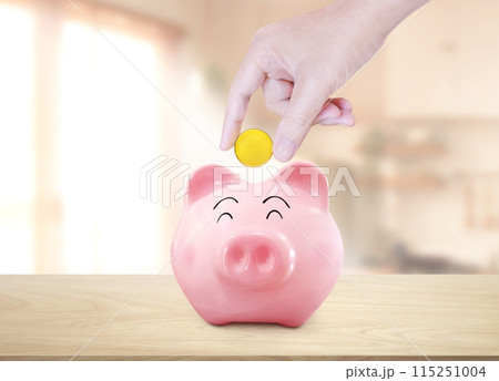 Hand puts gold coins into a piggy bank on a wooden table. Inside the room. 115251004