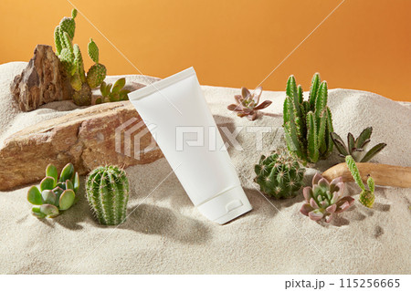 High angle view photo with an unlabeled cosmetic tube placed on sand and leaning on the rock beside, surrounded by some plants of desert, orange background. Mock up for advertising with space for text High angle view photo with an unlabeled cosmetic tube placed on sand and leaning on the rock beside, surrounded by some plants of desert, orange background. Mock up for advertising with space for text 115256665