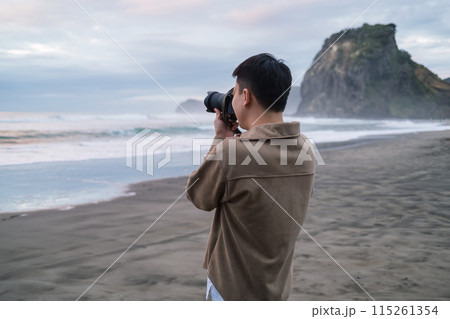 Male tourist is taking a photo with Lion Rock on Piha Beach in the evening at West of Auckland, New Zealand 115261354